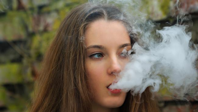 Vape teenager. Young pretty white girl in white cardigan smoking an electronic cigarette opposite destroyed brick wall on the street in the spring. Bad habit that is harmful to health.