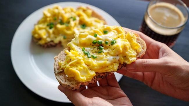 A cropped image of a woman's hand holding a piece of toasted bread with scrambled eggs on top, as she enjoys a healthy breakfast in the morning.