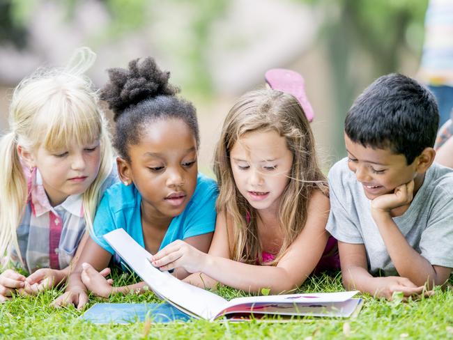 Three young girls and a boy are outdoors during summer. They are laying in the grass and reading a book together.