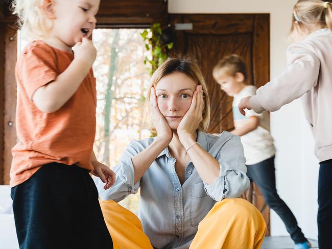 Frustrating woman alone struggling burnout with kids. Stressed out mother sitting on floor in middle of toys while children naughty running around her at room. Family home with chaos, mess.