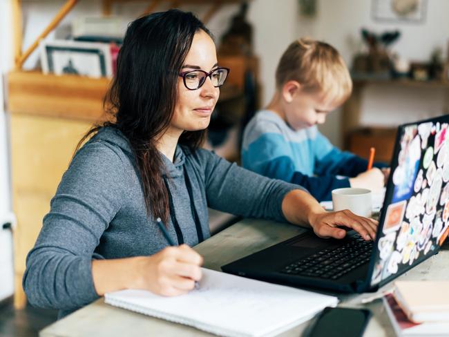 Concept of work from home and home family education. Mom and son are sitting at the desk. Business woman works on the Internet in a laptop, a child writes in a notebook.