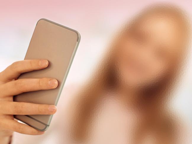 Helpful gadget. The focus being on the phone in the hands of a pretty fair-haired teenage girl standing in the room and taking a selfie while smiling