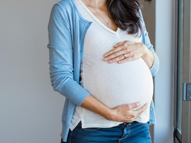 Portrait of mature pregnant woman at home looking outside the window. Young pregnant woman standing near window at home and thinking about the future. Smiling pregnant woman with hands on belly.