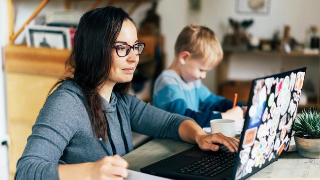 Concept of work from home and home family education. Mom and son are sitting at the desk. Business woman works on the Internet in a laptop, a child writes in a notebook.