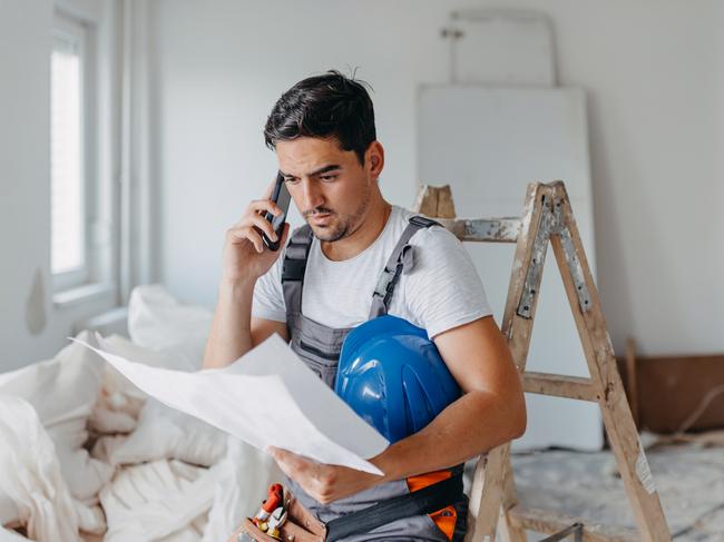 Developing Queensland - A young Caucasian male construction worker is having a phone call, while sitting down and holding the renovation plans.