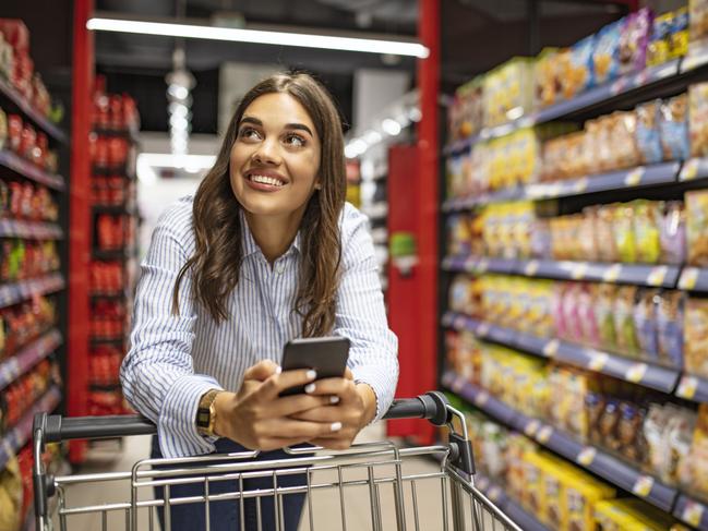 WOMAN MOBILE PHONE SUPERMARKET STOCK -  Smiling woman at supermarket. Happy woman at supermarket. Beautiful young woman shopping in a grocery store/supermarket. Shopping lists in app format Picture: Istock