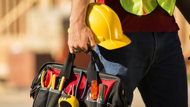 A construction worker busy working at a job site. He holds a tool box full of tools and a hard hat. Framed house, building in background. He is wearing a safety vest.