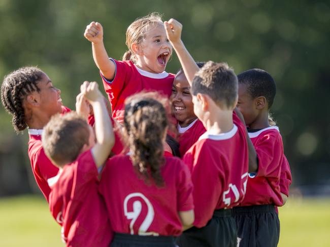 A multi-ethnic soccer team full of elementary age children are cheering together after winning their game. They have their arms raised up in the air victoriously.