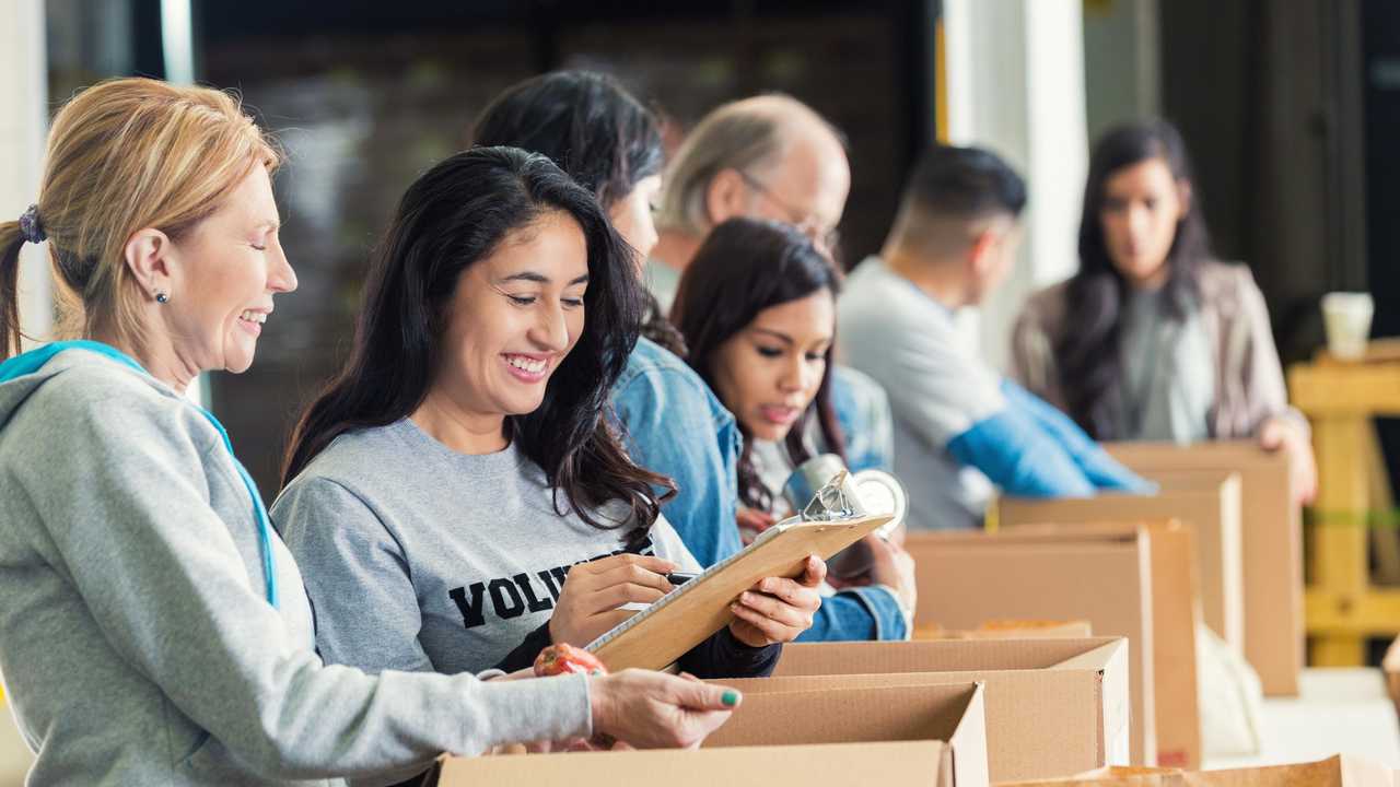 Mature adult Caucasian woman is standing with mid adult Hispanic woman as they pack cardboard boxees full of donated food in charity food bank. Other volunteers are lined up behind them, also sorting donated groceries into boxes. Hispanic woman is writing on checklist on clipboard. Picture: Steve Debenport