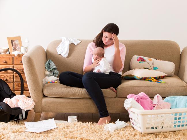 Mother with her baby on her knee. Her head is lowered as she sits on a sofa looking tired and stressed. The room looks untidy with clothes, nappies and other items scattered around the room. istock image