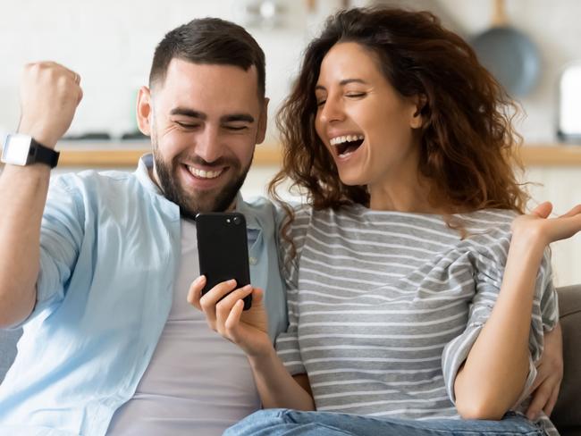 Excited millennial husband and wife relax on couch in kitchen feel euphoric win lottery online on cellphone, overjoyed young couple triumph celebrate read good unexpected news on smartphone gadget