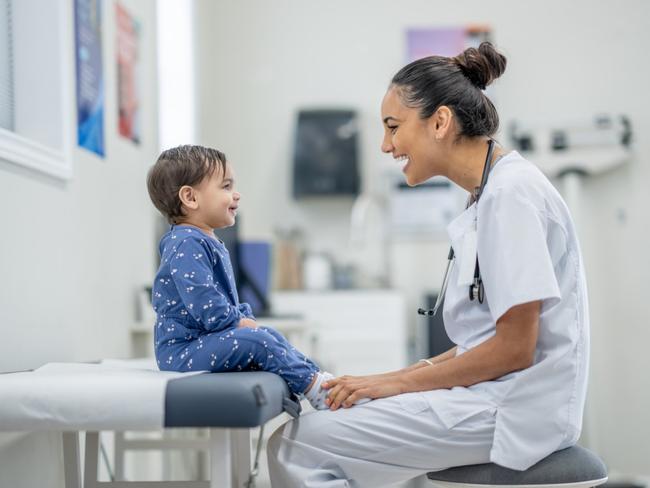 A sweet little toddler sits up on an exam table in a doctors office as her female doctor conducts a routine check-up.  The little girl is dressed casually and appears happy as her doctor engages with her.