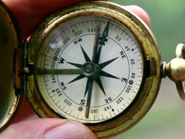 Old brass pocket compass held in fingertips with a blurred gravel path and woods in background. Direction, lost, bearings.