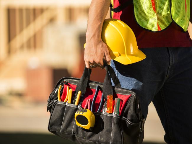 A construction worker busy working at a job site. He holds a tool box full of tools and a hard hat. Framed house, building in background. He is wearing a safety vest.