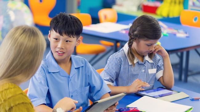 Teacher helping an Asian student in class. They are in a classroom, boy is wearing a school uniform. They are working at a desk. They are other ethnic backgrounds in shot