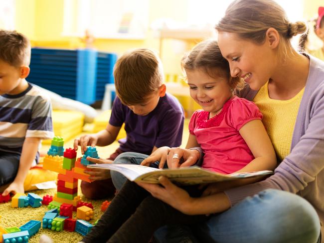 Happy little girl sitting in teacher's lap at preshool and reading a book while other children are playing.