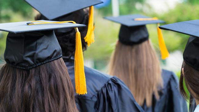 Group of students wearing graduation caps and gowns are walking on campus lawn. Young men and women are graduating from high school or college. Photo is of backs of students heads as they walk to receive their diplomas. Black caps have gold tassles.