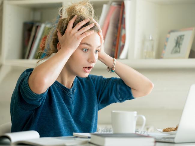 money mistakes - generic woman shocked, stressed... Portrait of an attractive woman at the table with cup and laptop, book, notebook on it, grabbing her head. Bookshelf at the background, concept photo