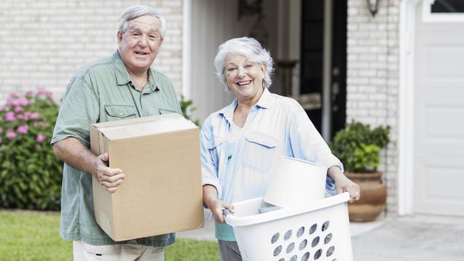 A senior couple relocating, downsizing to a smaller home. They are carrying a box and laundry basket into their new house, looking at the camera and smiling. They are in their 60s.