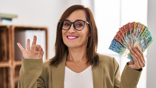 Middle age woman working at consultation office holding australian dollars doing ok sign with fingers; women female investing money generic