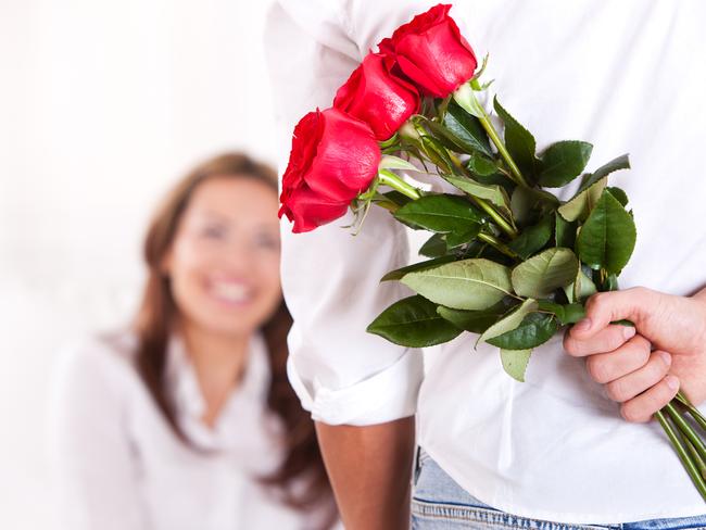 Close-up of young man holding bouquet of red roses behind back. Defocused young woman in the background.