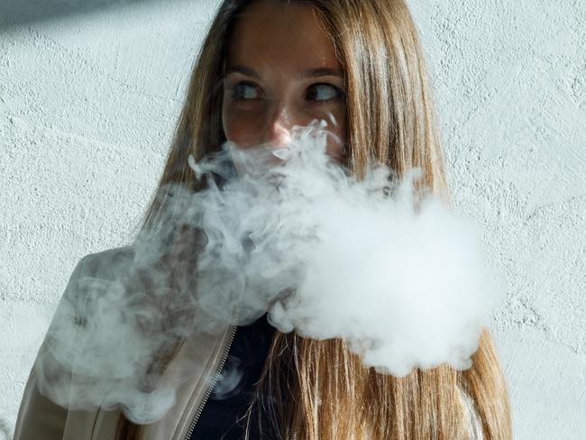 Vape teenager. Young cute girl in  casual clothes smokes an electronic cigarette near the wall outdoors in summer day. Bad habit that is harmful to health. Vaping activity.