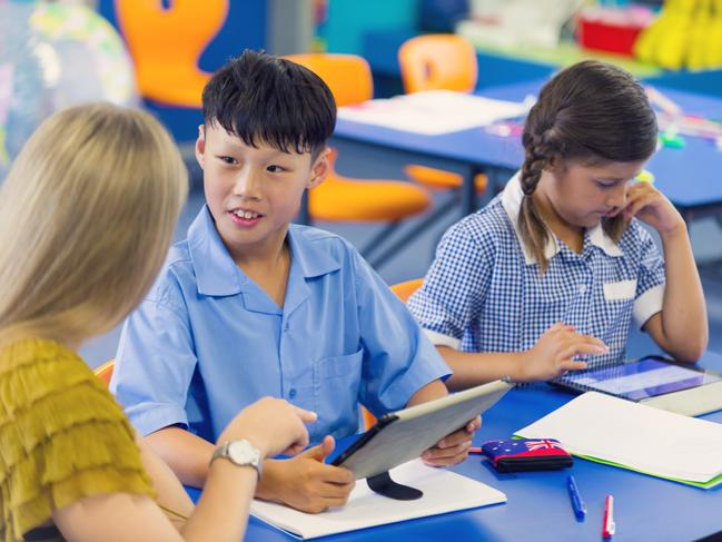 Teacher helping an Asian student in class. They are in a classroom, boy is wearing a school uniform. They are working at a desk. They are other ethnic backgrounds in shot