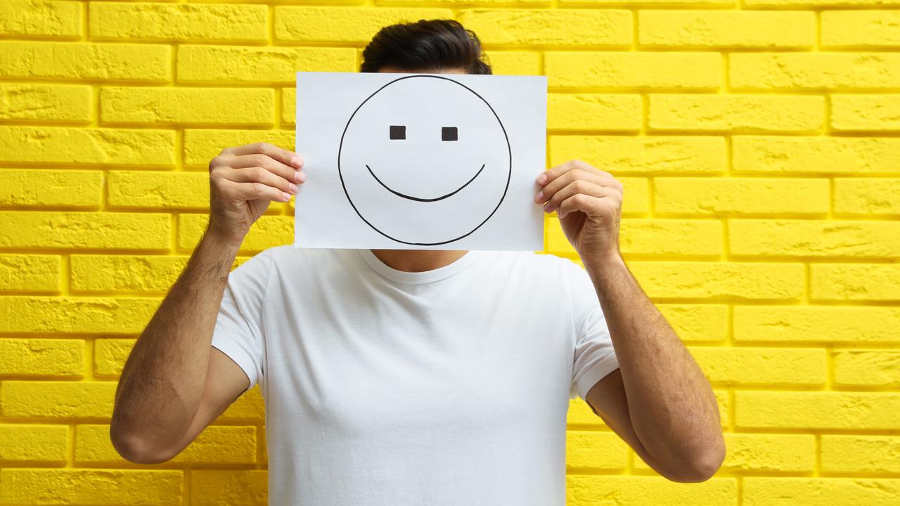 Man hiding emotions using card with drawn smiling face near yellow brick wall