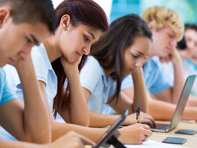 Row of private high school students work on assignment in class. They are writing or using laptops or digital tablets. They are concentrating as they study. They are wearing school uniforms.