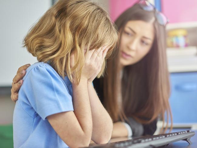 a primary school girl is sitting at her computer desk in school and crying into her hands. Her young teacher looks on in the background, puts her arm around her and gives support.