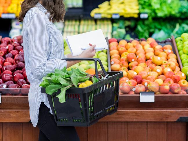 Young woman carries a shopping basket filled with fresh produce. She is shopping for fresh fruit and vegetables in a grocery store.