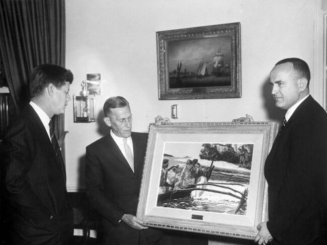 President John F. Kennedy visits with A.R. "Reg" Evans (left). Evans is holding a painting that shows Kennedy being helped into a canoe.