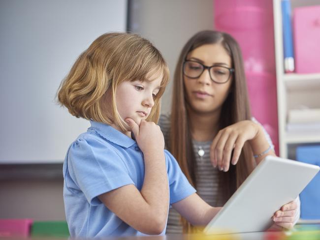 a young classroom assistant  listens as her young student explains what she is reading on a digital tablet . The little girl is having a think with her hand on her chin . Picture: Istock