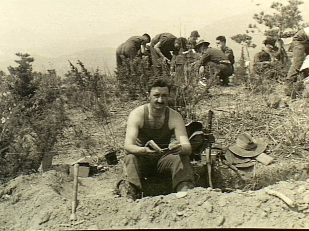 My country, write or wrong ... Corporal G.F. “Fibber” McGee, a member of the Machine Gun Platoon, 3rd Battalion, The Royal Australian Regiment, sits on the edge of a trench or dugout and reads a book in Korea, while comrades unload ration boxes. Picture: Australian War Memorial