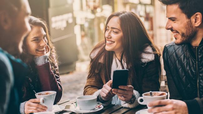 Laughing young people looking at phone and laughing while sitting in cafe outdoors, holding cups.