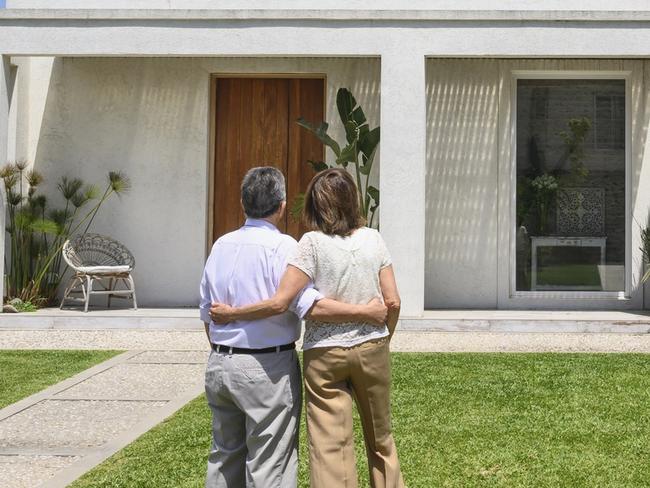 Rear view of senior couple standing in front yard with arms around each other and admiring two-story Buenos Aires family home.