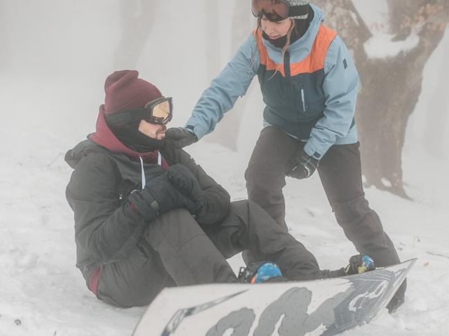 A male snowboarder sits on the snow after a fall, while another snowboarder approaches him and offers help