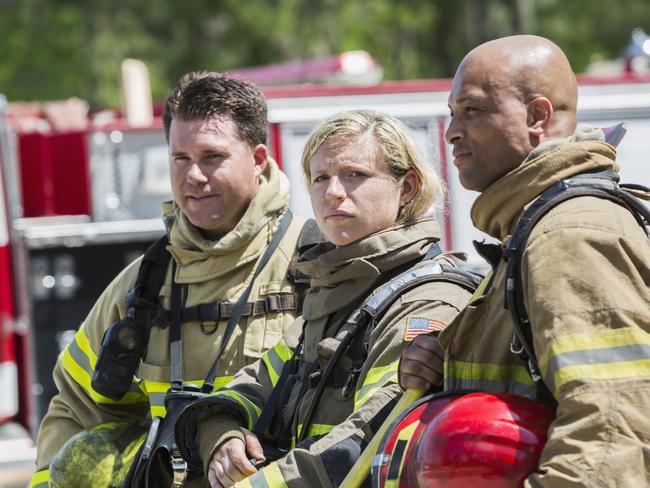 A female firefighter standing with two males colleagues in front of a fire engine.  She is looking at the camera with a serious expression.  They are wearing tan protective suits with yellow reflective stripes, carrying helmets and oxygen.