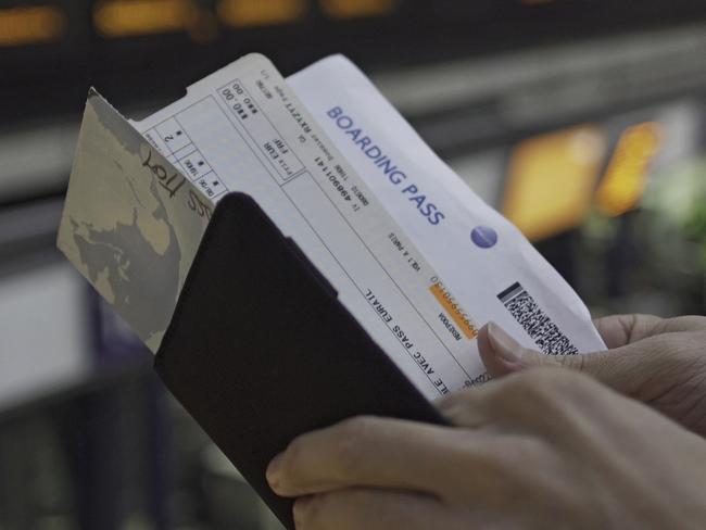 A pair of man's hands checks his travel documents and the time on his wristwatch. In the background is a large, out of focus, electronic departures board.