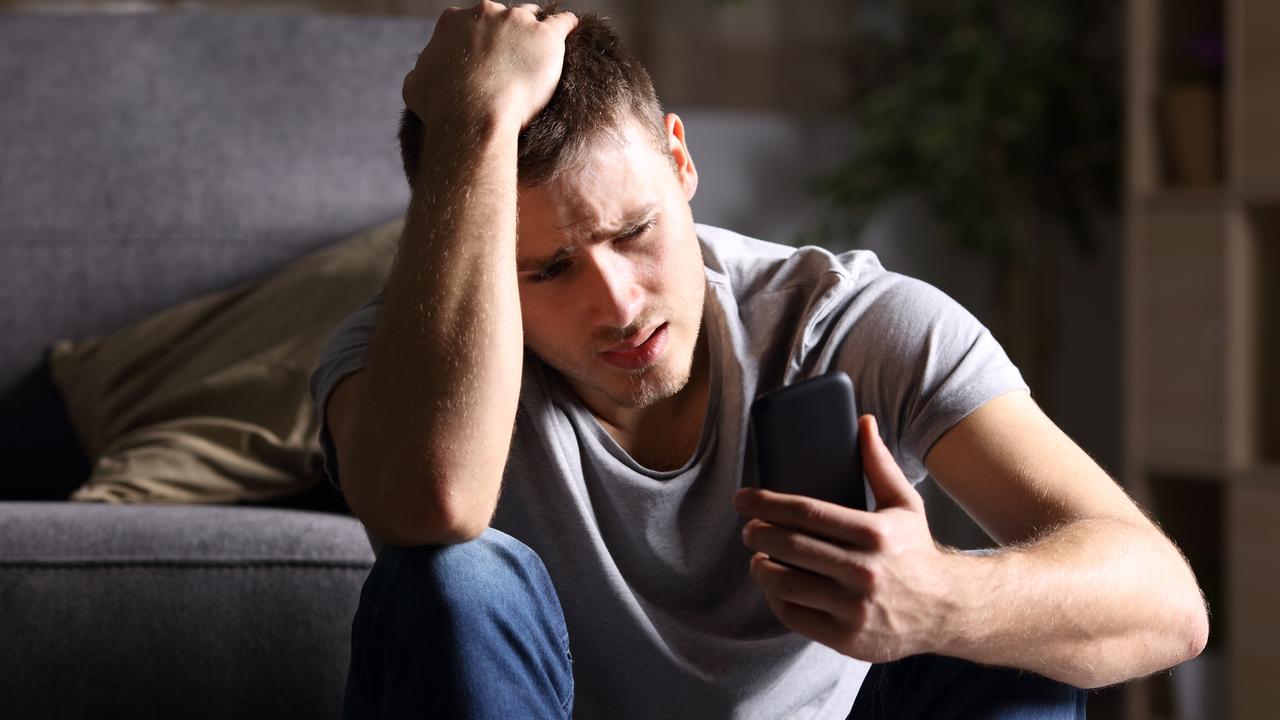 Single sad man checking mobile phone sitting on the floor in the living room at home with a dark background