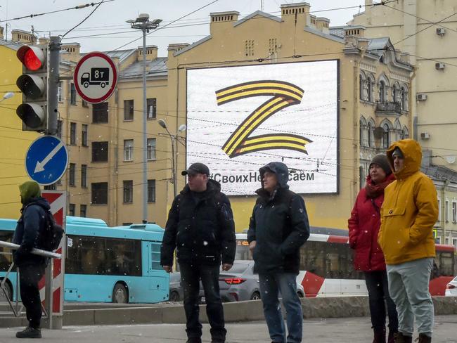 Pedestrians cross a street in front of a billboard displaying the symbol “Z” in the colours of the ribbon of Saint George and a slogan reading: 