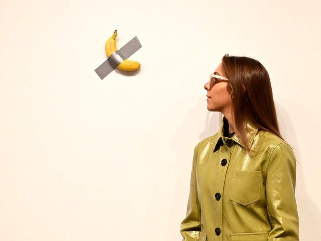 A woman has her photo taken beside a banana duct taped to the wall, an artwork titled 'Comedian' by Italian artist Maurizio Cattelan, during a preview of the National Gallery of Victoria's (NGV) Triennial 2023, an exhibition featuring work by over 120 contemporary artists, designers and collectives, in Melbourne on December 1, 2023. (Photo by William WEST / AFP) / RESTRICTED TO EDITORIAL USE - MANDATORY MENTION OF THE ARTIST UPON PUBLICATION - TO ILLUSTRATE THE EVENT AS SPECIFIED IN THE CAPTION