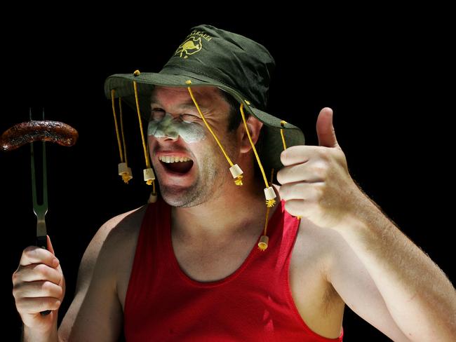 An Aussie man in a cork hat winks at the camera holding up a sausage on a fork in one hand and giving the thumbs up gesture with the other