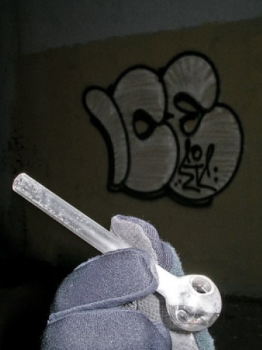 A typical crystal meth pipe is held up in front of graffiti glorifying drug use.