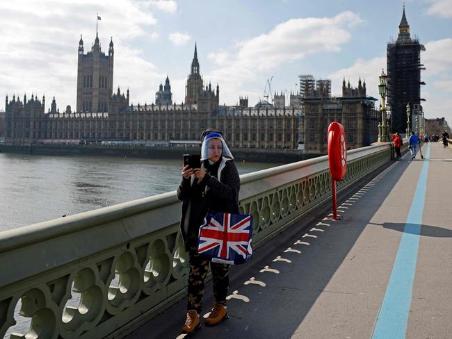 A pedestrian wearing a face shield due to Covid-19, stands with a Union flag-themed bag as they make a video call in front of the Palace of Westminster.