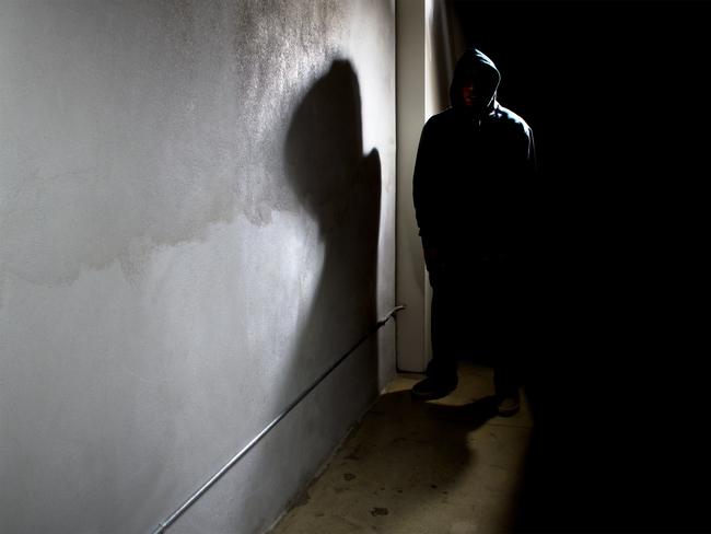 Photo of a hooded criminal stalking in the shadows of a dark street alley.  The hooded man is a silhouette and hiding in the dark.  The man is a criminal waiting to ambush victims.  The concrete walls provide copyspace.The photo depicts crime.  - istock