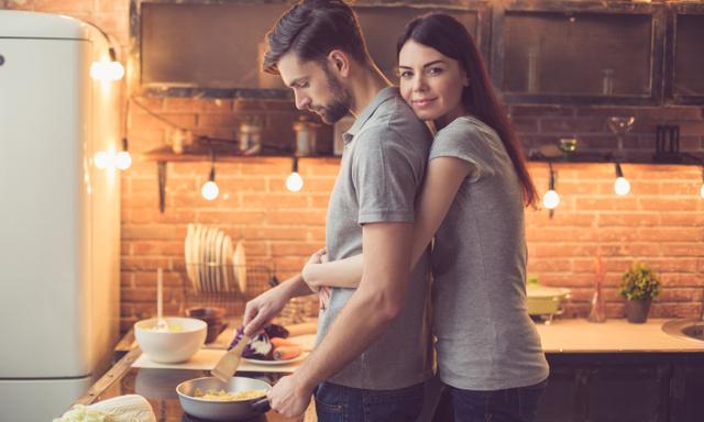 Young beautiful couple in kitchen. Family of two preparing food. Woman looking at camera and hugging man while he making delicious pasta. Nice loft interior with light bulbs