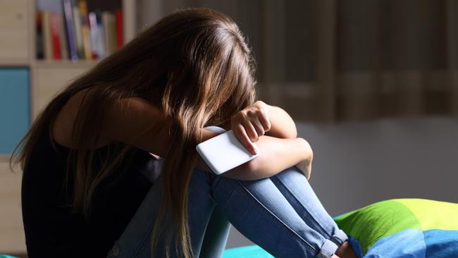 Single sad teen holding a mobile phone lamenting sitting on the bed in her bedroom with a dark light in the background