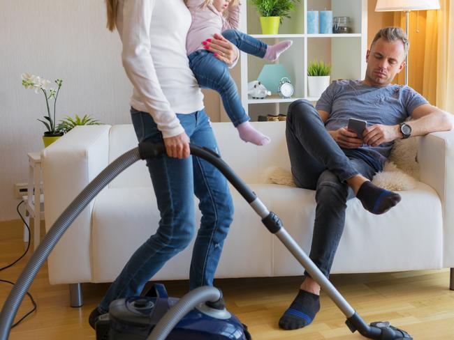 Woman with small child in her hands doing housekeeping while man sitting in couch and relaxing.