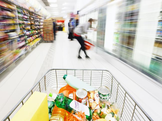 View over filled shopping cart racing down a supermarket aisle towards out-of-focus  shoppers, showing extreme, multicolored motion blur.  The product labels in the cart are made by the photographer